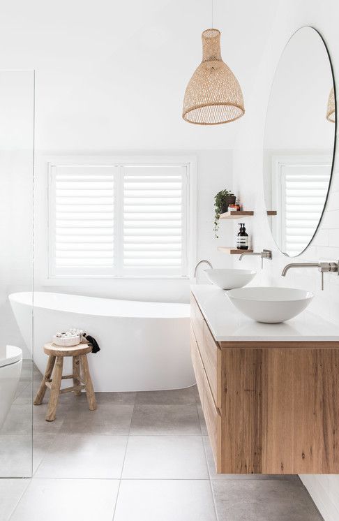 a modern white coastal bathroom with large format tiles, a timber vanity, an oval tub, a wooden stool and a woven pendant lamp
