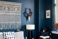 a moody boho bedroom with navy walls, black chairs, a macrame hanging, a woven lamp and printed bedding