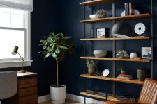 a moody midnight blue home office with industrial furniture, a leather chair and ottoman, a shelving unit and a pendant lamp