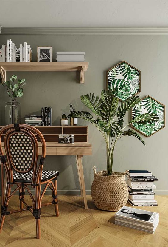 a moody tropical home office with an olive green wall, tropical artworks, a monstera in a basket and a rattan chair