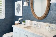 a nautical bathroom with blue grasscloth wallpaper, a neutral vanity, a mirror covered with rope, some sea-inspired decor and white appliances
