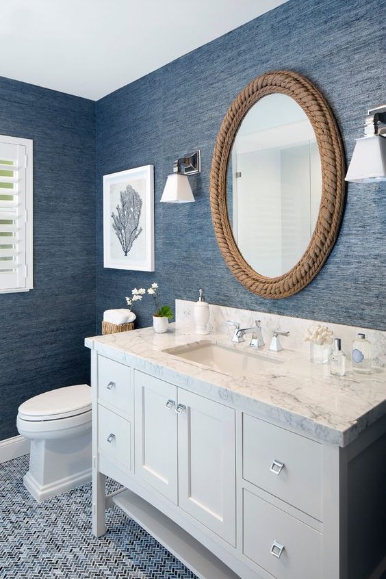 a nautical bathroom with blue grasscloth wallpaper, a neutral vanity, a mirror covered with rope, some sea-inspired decor and white appliances