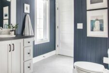 a nautical bathroom with navy shiplap walls, a white vanity, some artwork and white appliances, black sconces