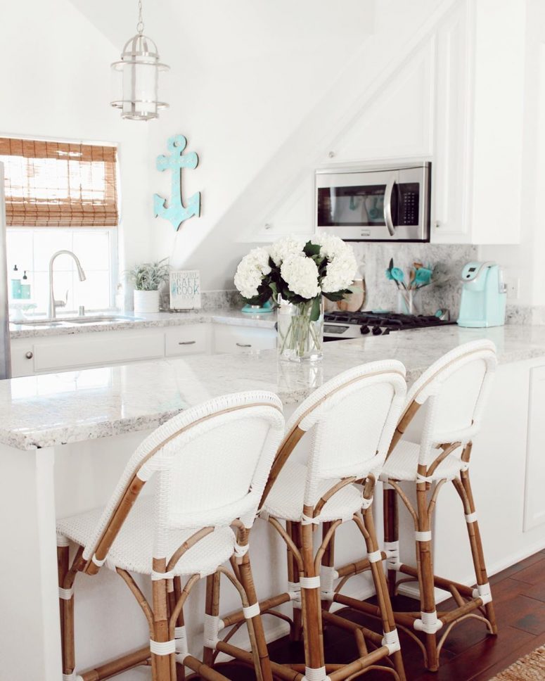 a neutral coastal kitchen with white cabinetry, a grey stone countertop, woven shades, a light blue anchor and rattan chairs