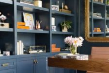 a preppy navy home office with built-in storage units, a wood and acryl desk, a gold framed mirror and a sunburst chandelier