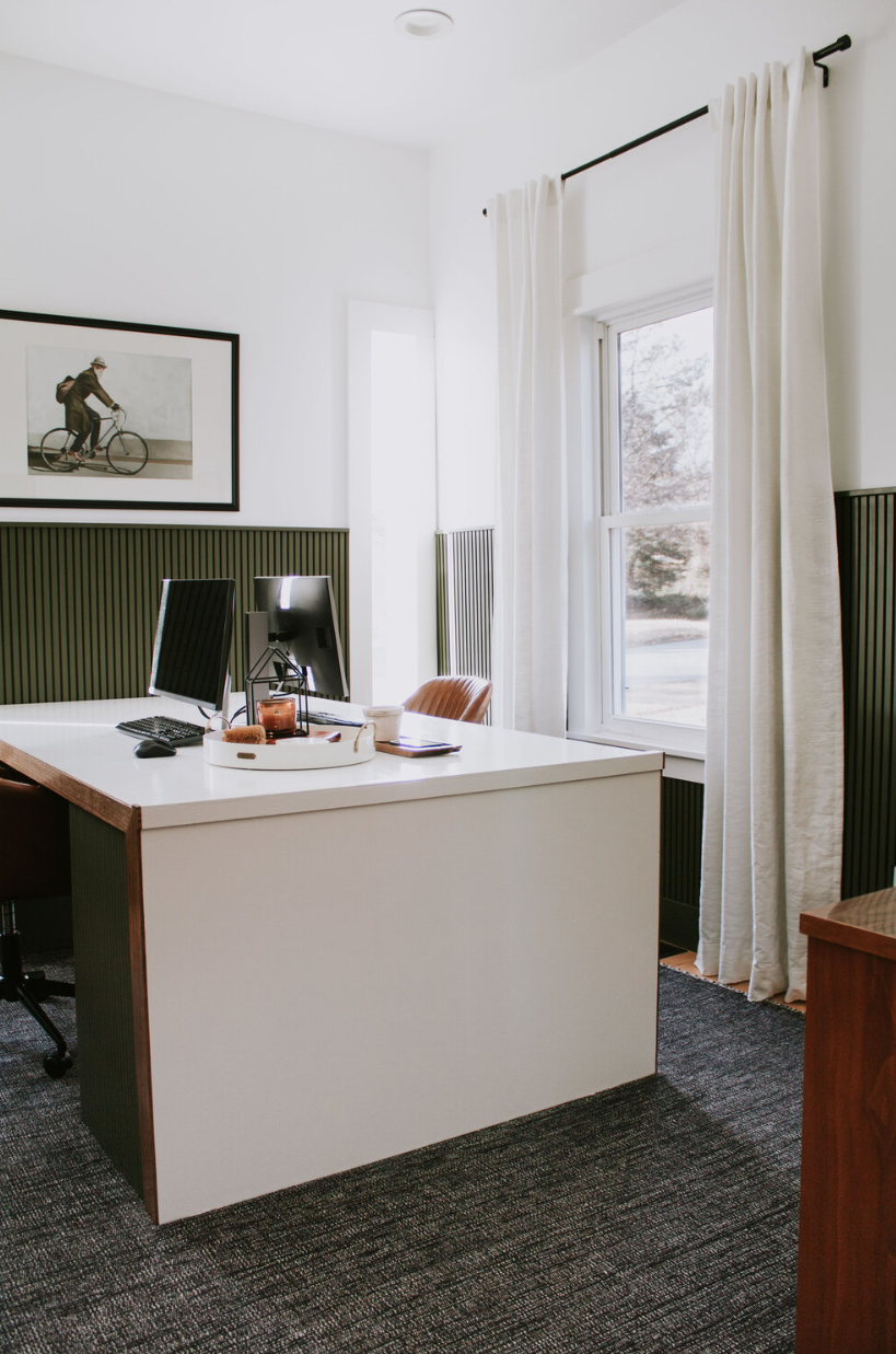 a refined modern double home office with sage green wooden planks on the walls, a chic color block desk, leather chairs and a statement artworkk