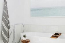 a serene bathroom with large format marble tiles, an oval tub, a coastal artwork, a wooden stool and a striped towel