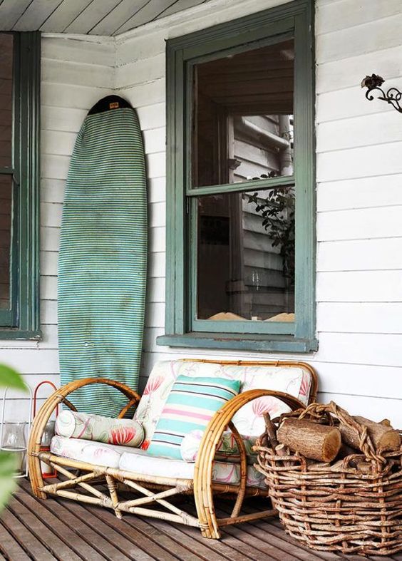 a simple and lovely beach porch with a large rattan chair with colorful textiles, a turquoise fabric covered surf board and a basket with firewood