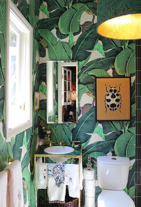 a small and fun powder room with banana leaf wallpaper, a small green and gold vanity and a bright artwork