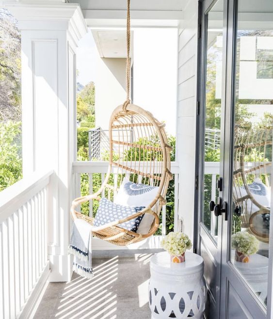 a small neutral coastal porch with a hanging rattan chair with blue pillows and a white side table plus a view