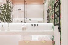 a spacious tropical bathroom with tropical wallpaper, a blush cabinet with gold handles, a pink ottoman and a large mirror