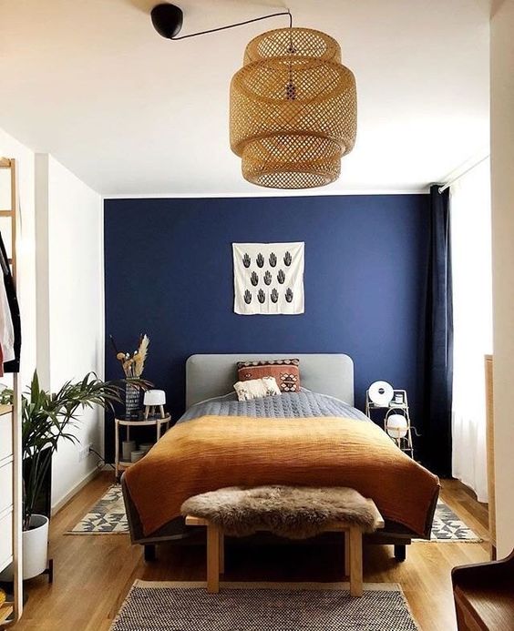 a stylish bedroom with a navy accent wall, a grey bed with printed bedding, woven lamp, mismatching nightstands and potted greenery