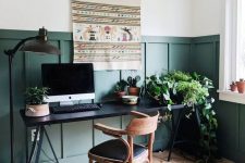 a stylish boho modern home office with hunter grene paneling, a black desk, a wooden chair and a folksy hanging on the wall
