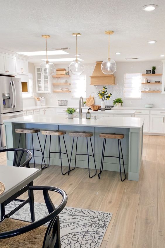 a stylish coastal farmhouse kitchen with white cabinets, a light blue island, wooden shelves and a hood, weathered wood stools