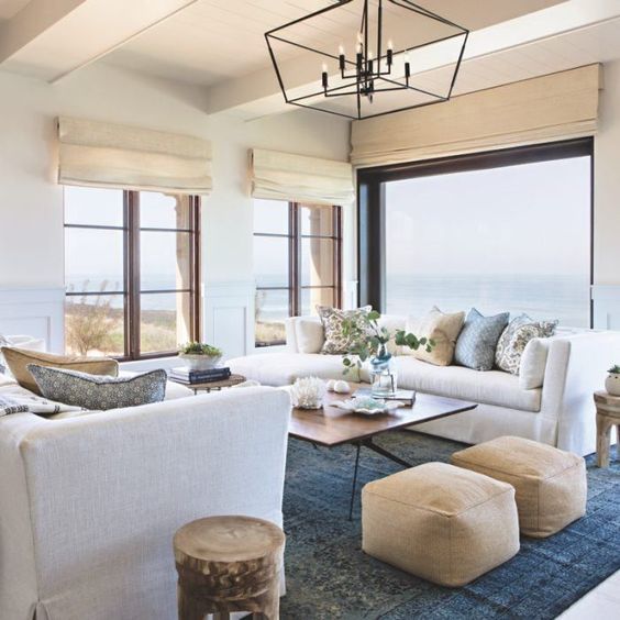 a stylish coastal living room with large windows including a panoramic one, with shades, leather ottomans and touches of blue