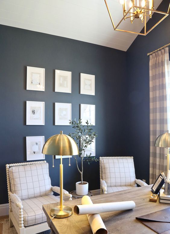 a stylish farmhouse home office with navy walls, a wooden desk, windowpane chairs, a chic chandelier and brass lamps