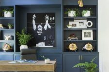 a stylish home office with a blue storage unit that takes one wall, a wooden desk, a leather chair and touches of greenery and gold