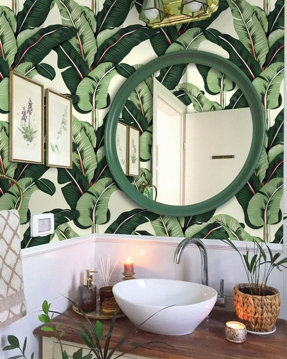 a stylish tropical powder room with banana leaf wallpaper, a grene framed mirror, a white bowl sink and some greenery