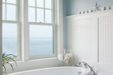 a traditional sea-inspired bathroom done in white and blue, with an oval sunken tub and a gorgeous view to the sea