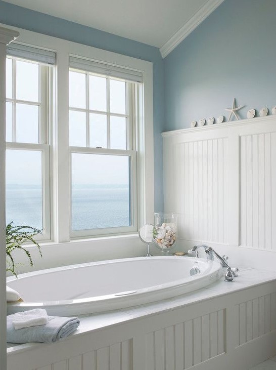 a traditional sea-inspired bathroom done in white and blue, with an oval sunken tub and a gorgeous view to the sea
