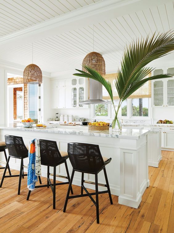 a tropical kitchen in white, with white cabinets and stone countertops, rattan pendant lamps and black stools