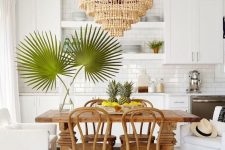 a tropical kitchen with white cabinets, a subway tile backsplash, a wooden table and chairs plus a bead chandelier and tropical leaves