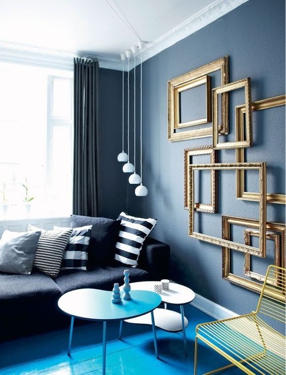a unique living room with a bright blue floor, grey walls, a black sofa, monochromatic pillows and gold frames on the wall