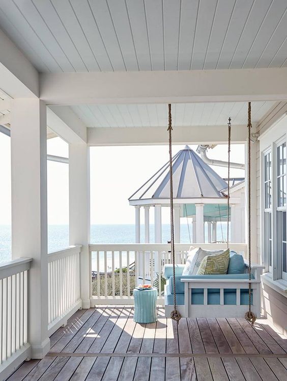 a very simple beach porch with a white hanging daybed with blue upholstery and a blue side table plus a view