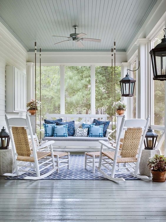 a vintage coastal porch with white furniture, blue pillows, a blue printed rug, potted greenery and candle lanterns