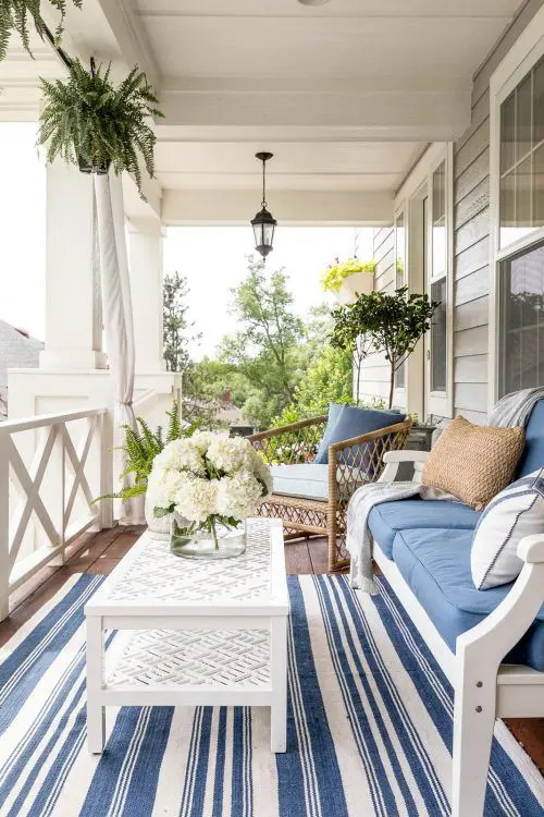 a vintage coastal porch with white wooden and rattan furniture, blue and white textiles, a white table and a striped rug