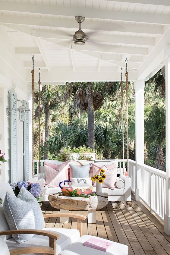 a welcoming beach porch with a hanging sofa, a comfy lounger, pastel textiles, a wood table and blooms