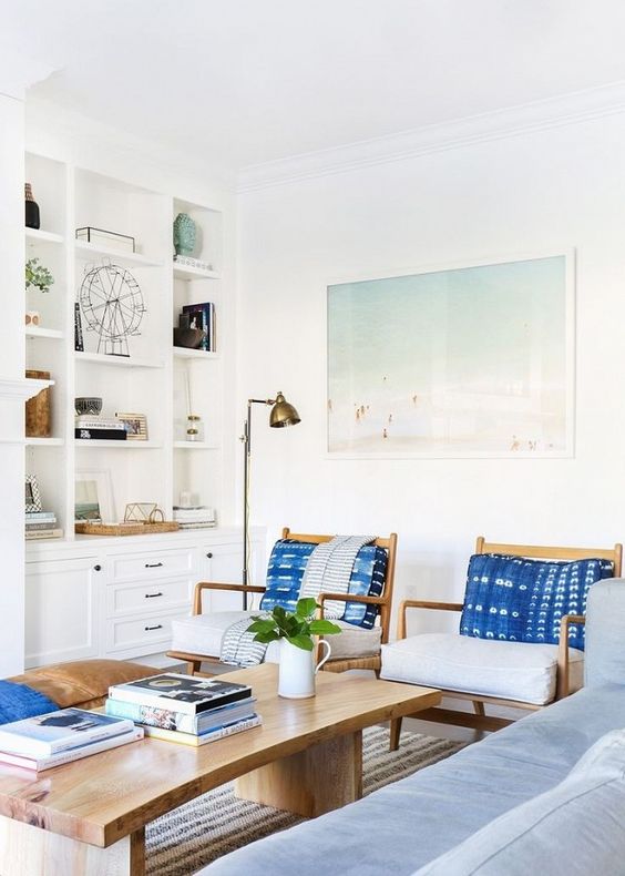 a welcoming coastal living room with blue furniture, indigo pillows, a wooden table, a leather ottoman and a beach artwork