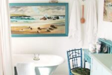 a whimsical coastal bathroom with a blue clawfoot tub, a vanity, a chair and a makeup table, some artwork and towels