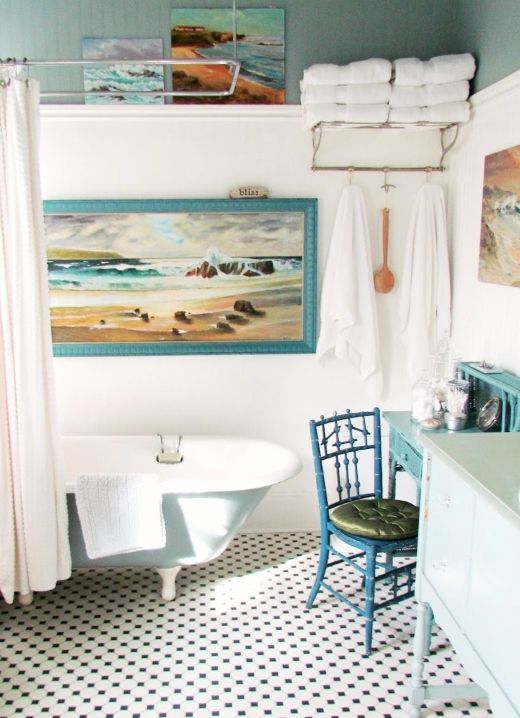 a whimsical coastal bathroom with a blue clawfoot tub, a vanity, a chair and a makeup table, some artwork and towels