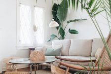 an airy living room with white walls, a neutral sofa, rattan chairs, a glass coffee table and potted plants