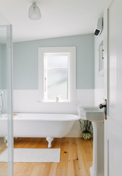 an airy retro inspired coastal bathroom with color block pale blue and white walls, a clawfoot tub, a free standing sink and neutral linens