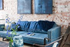 an eclectic living space with an exposed brick wall, a classic blue sofa, indigo artworks and pillows and a black rug