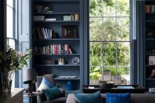 an elegant blue living room with blue walls and built-in shelves, grey and brown furniture, potted plants and a cluster chandelier