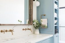 an enchanting light blue coastal bathroom with lovely vintage furniture, white marble countertops, a large wood framed mirror and touches of brass