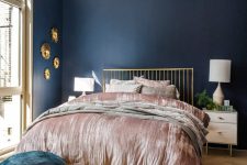 an exquisite bedroom with navy walls, a gold bed, neutral nightstands, navy and gold ottomans and a pretty gallery wall