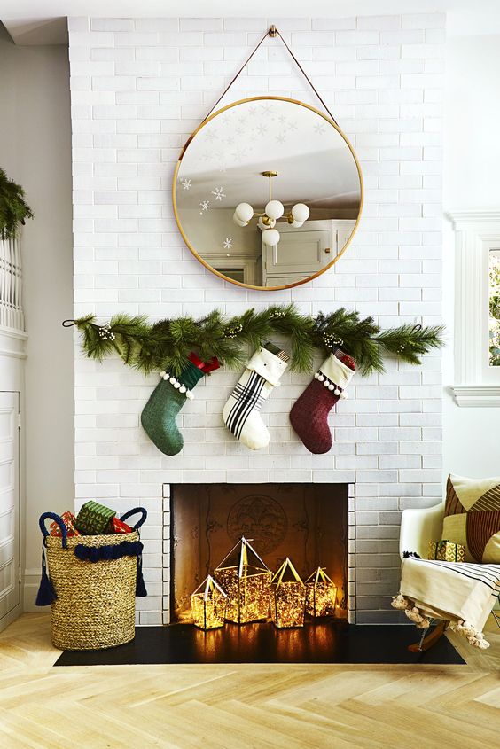 a Christmas fireplace with candle lanterns and lights, evergreens and stockings over the fireplace and a basket with gifts