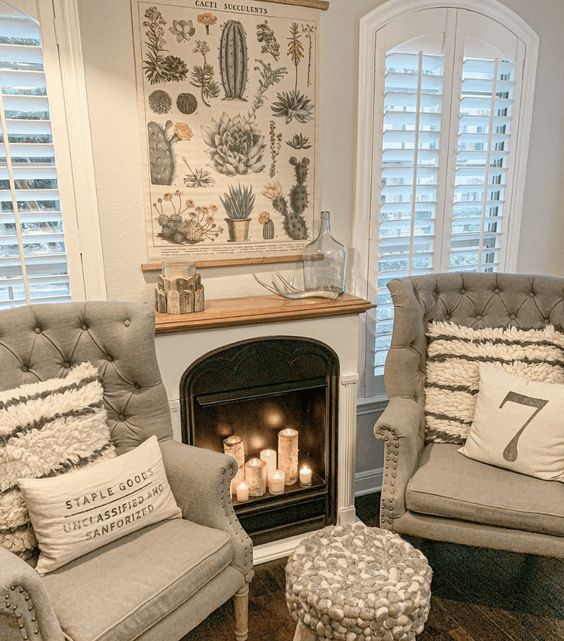 A built in non working fireplace with pillar candles, grey chairs with pillows and a pouf