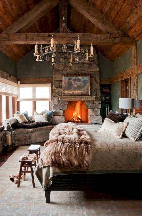 a cabin bedroom with a rough stone fireplace and a bench, a catchy chandelier, wooden items and a cozy linens