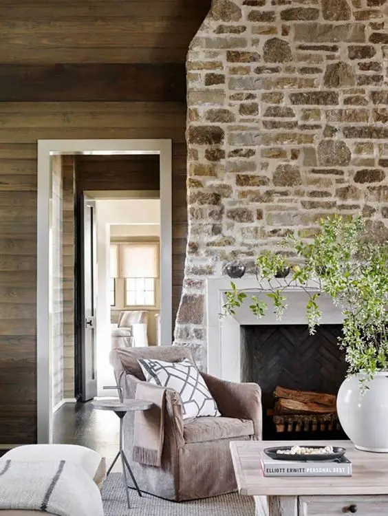 a chalet living room with a stone fireplace, some neutral seating furniture, a low coffee table and greenery
