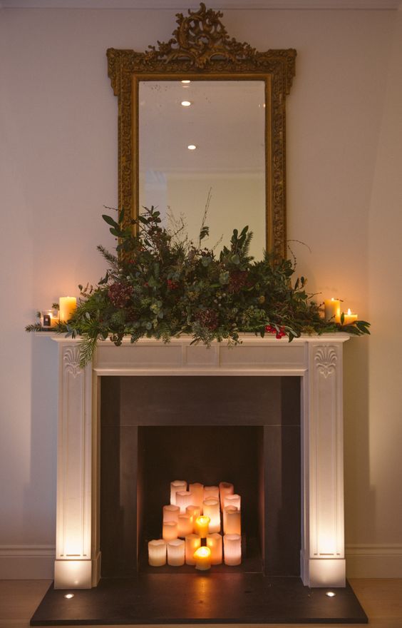 a chic Christmas fireplace with lots of candles and an evergreen and pinecone arrangement plus a mirror on top