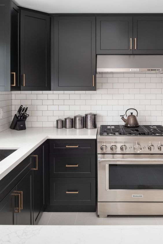 a contrasting kitchen with black cabinets, a white subway tile backsplash and white stone countertops plus brass handles