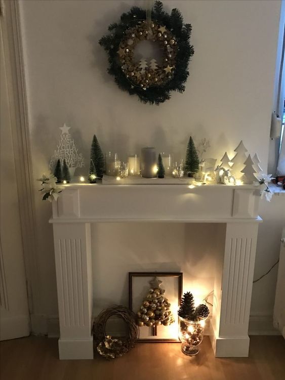 a faux fireplace with a Christmas tree art, a vase with pinecones, a Christmas arrangement on the mantel