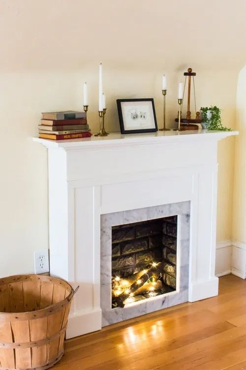 a faux fireplace with logs and lights, with a mantel styled with books, candles and greenery is cozy and cool