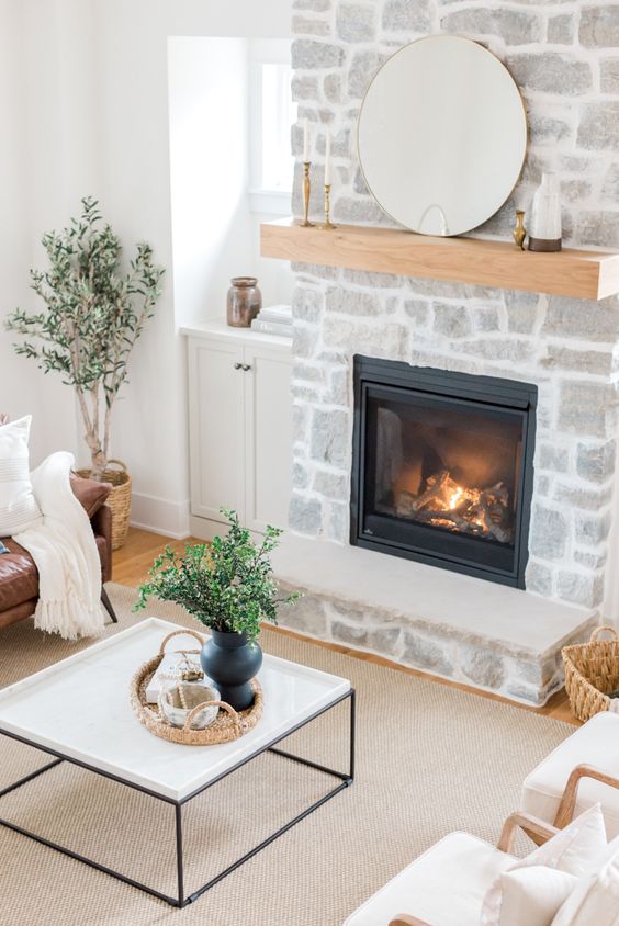 a fireplace clad with stone, with a mantel, a leather sofa, a coffee table and neutral chairs, potted greenery