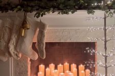a fireplace with pillar candles, branches, a lit up tree, white stockings and greenery on the mantel